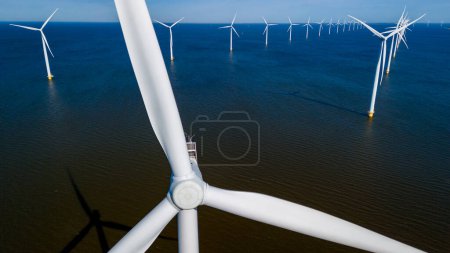 Foto de Un grupo de turbinas de molino de viento en las aguas del océano, sus cuchillas girando en la suave brisa primaveral de Flevoland, Países Bajos. vista aérea de drones de turbinas de molino de viento energía verde en el océano - Imagen libre de derechos