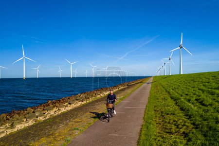 Foto de Un hombre en bicicleta enérgicamente por un camino bordeado de turbinas eólicas en los Países Bajos Flevoland durante la vibrante temporada de primavera. hombres en bicicleta eléctrica con turbinas de molino de viento en el fondo - Imagen libre de derechos