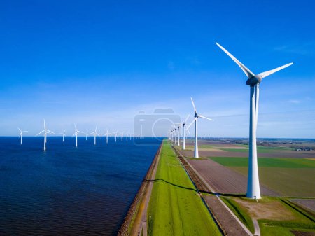 Photo for A serene scene of a row of wind turbines standing tall next to a calm body of water in Flevoland, the Netherlands, during the vibrant season of Spring. green energy in the ocean - Royalty Free Image