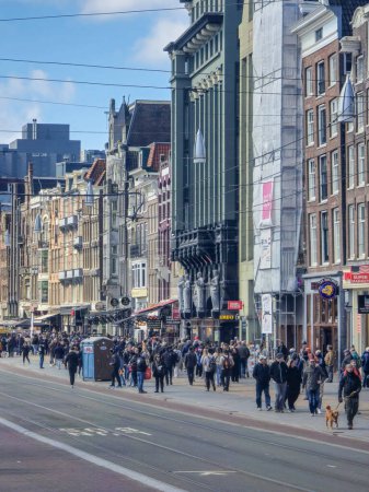 Téléchargez les photos : Amsterdam Pays-Bas 21 avril 2024 Un groupe diversifié de personnes marche dans une bousculade animée dans une rue animée de la ville flanquée de magasins, sous un ciel bleu clair. - en image libre de droit