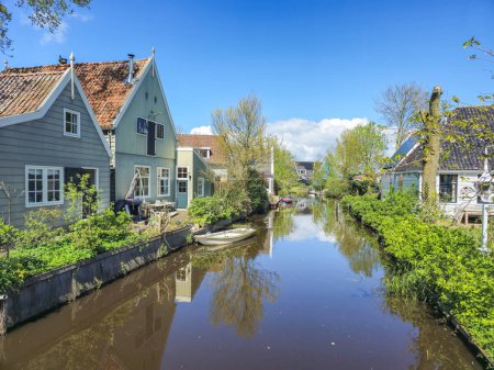 A tranquil river flows peacefully through a lush, green countryside with vibrant foliage and rolling hills under a clear blue sky. Broek in Waterland Netherlands 