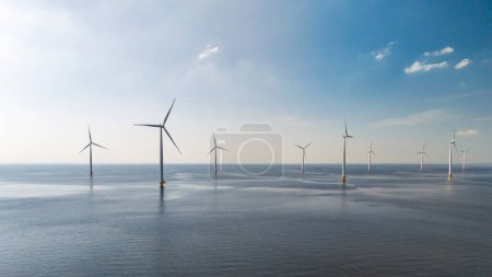 Foto de Un grupo de molinos de viento virando con gracia sus cuchillas contra el fondo del vasto océano en un día soleado en Flevoland, Holanda. Turbinas eólicas en el mar, transición energética verde en Europa - Imagen libre de derechos
