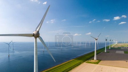 Photo for A row of wind turbines stands tall next to a serene body of water, their blades spinning gracefully in the wind, generating clean energy. - Royalty Free Image