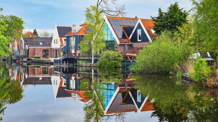 rangée de charmantes maisons nichées à côté d'un plan d'eau tranquille, reflétant le ciel bleu clair et la verdure luxuriante.façades en bois et vieilles maisons à Broek dans Waterland aux Pays-Bas