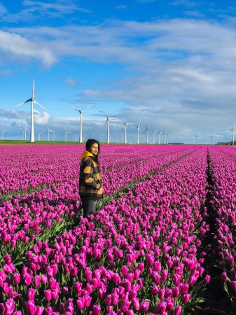 Eine Asiatin steht stolz auf einem Feld mit leuchtend violetten Blumen, umgeben von der Schönheit der Natur, die an einem sonnigen Tag in voller Blüte steht. Asiatische Frauen mit Windkraftanlagen im Hintergrund