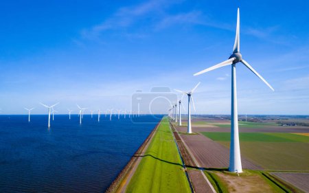 Foto de Una fila de aerogeneradores se encuentra alto junto a un tranquilo cuerpo de agua en los Países Bajos Flevoland durante springtime.drone vista aérea de aerogeneradores energía verde en el océano - Imagen libre de derechos