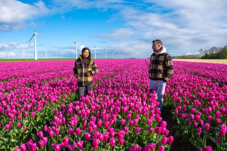 A couple embraces a vast field of vibrant purple tulips, under the watchful gaze of towering windmill turbines in the Netherlands in Spring. A diverse couple of Asian women and caucasian men