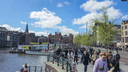 Téléchargez les photos : Amsterdam Pays-Bas 21 avril 2024, Un groupe diversifié de personnes se promènent dans une rue charmante au bord d'une rivière paisible, admirant le paysage et bavardant animément. - en image libre de droit