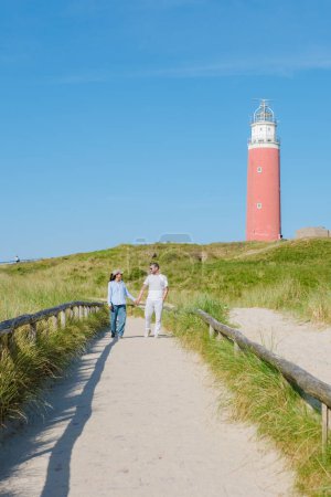 Eine friedliche Szene spielt sich ab, als zwei Personen auf einem Pfad in der Nähe eines charmanten Leuchtturms auf Texel, Niederlande, spazieren. Ein Paar Mann und Frau am berühmten roten Leuchtturm von Texel