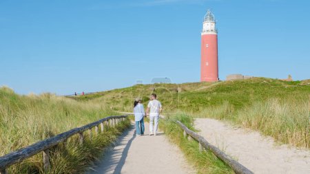 Ein Paar spaziert gemütlich auf einem gewundenen Pfad in der Nähe des ikonischen Leuchtturms von Texel und genießt an einem friedlichen Tag die malerische Aussicht auf die Küste. Mann und Frau am berühmten roten Leuchtturm von Texel