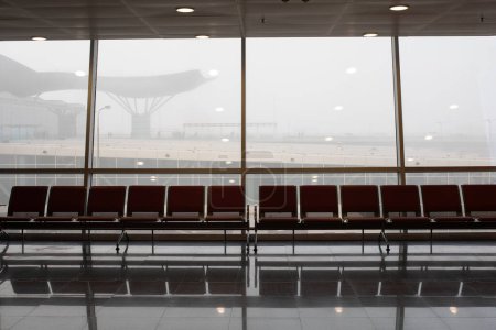 Foto de Asientos vacíos en la terminal del aeropuerto. La niebla gruesa es visible fuera de la ventana. Concepto de mal tiempo, cancelaciones de vuelos, retrasos de vuelos. - Imagen libre de derechos