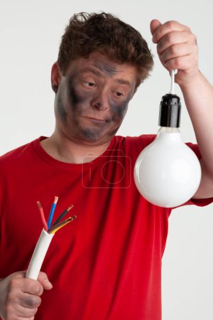 Téléchargez les photos : L'homme tient une lampe dans une main et un câble dans une autre. Son visage est fumé de suie noire à la suite d'un court-circuit. Il porte un t-shirt rouge comme avertissement de risque réparation non autorisée - en image libre de droit