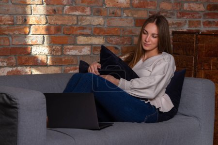 Foto de Mujer sonriente en un suéter blanco sentada en el sofá, sosteniendo una almohada y mirando a un ordenador portátil - Imagen libre de derechos