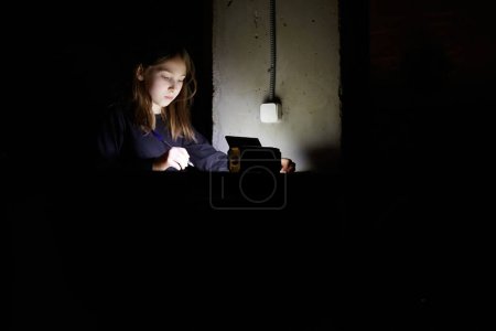 Téléchargez les photos : La fille apprend des leçons dans le noir. Un enfant travaille à l'école lorsqu'il n'y a pas d'électricité à la maison - en image libre de droit