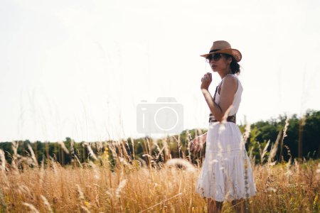 Téléchargez les photos : Portrait de jeune femme marchant parmi les hautes herbes dans la prairie d'été portant un chapeau de paille et robe de lin profitant de la nature. Harmonie et équilibre. - en image libre de droit