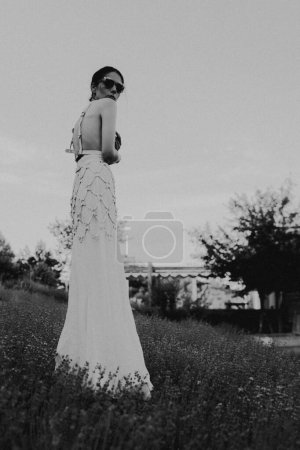 Foto de Hermosa mujer morena joven en vestido blanco y gafas de sol posando en un campo de lavanda en la noche de verano. Concepto de belleza - Imagen libre de derechos