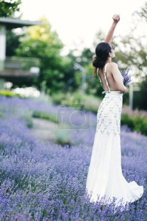 Téléchargez les photos : Belle jeune femme brune en robe blanche et lunettes de soleil posant dans un champ de lavande le soir d'été. Concept de beauté - en image libre de droit