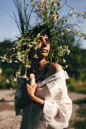 Foto de Una joven en un vestido blanco y una corona de margaritas sostiene en sus manos un pequeño chiken, sobre el fondo del bosque, la luz del atardecer. Concepto de libertad - Imagen libre de derechos