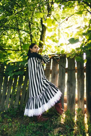 Téléchargez les photos : Élégante jeune femme indienne en robe noire et blanche sur fond d'arbres verts et de clôtures en bois vieilli. Femme Boho se détendre dans la campagne, style de vie lent simple. Image atmosphérique. - en image libre de droit