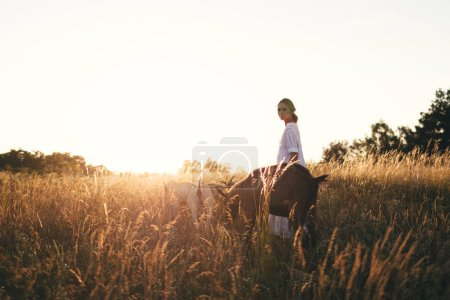 Foto de Mujer joven en vestido blanco está caminando con cabras en el prado al atardecer. Atractiva agricultora alimentando a sus cabras en su pequeña granja orgánica. - Imagen libre de derechos