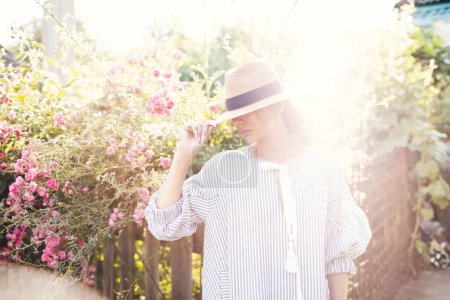 Téléchargez les photos : Romantique élégamment habillé jeune adulte belle femme se détendre dans un parc de campagne regardant loin. Modèle blond de style parisien dans une robe blanche transparente et chapeau de paille marchant contre du jardin. - en image libre de droit