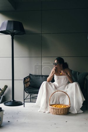 Téléchargez les photos : Portrait d'une belle femme indienne en robe blanche et lunettes de soleil assises sur le canapé avec un panier de cerise douce jaune. - en image libre de droit