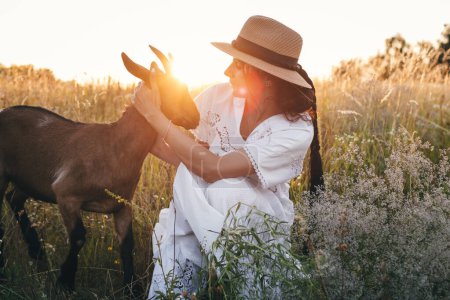 Téléchargez les photos : Une jeune femme sur le terrain marche avec des chèvres. La fille et les chèvres dans la prairie en été. L'amour pour les animaux. Une ferme de chèvres. Les animaux. Une femme heureuse avec un animal. Bonté et amour pour les animaux. Embrasse un animal - en image libre de droit
