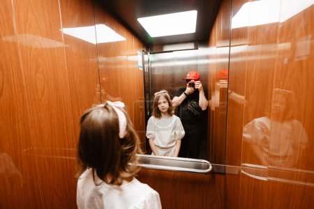 Téléchargez les photos : Père heureux faisant la photo de sa fille en utilisant un appareil photo professionnel dans l'ascenseur. - en image libre de droit