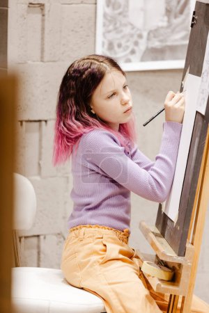 Photo for Girl 11 years old craftswoman are painting on canvas in studio standing in front of easel. Portrait of a girl painting during an art class - Royalty Free Image