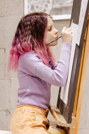 Photo for Girl 11 years old craftswoman are painting on canvas in studio standing in front of easel. Portrait of a girl painting during an art class - Royalty Free Image