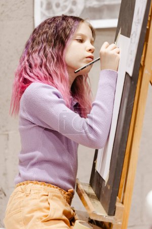 Foto de Chica 11 años de edad artesana están pintando sobre lienzo en el estudio de pie delante del caballete. Retrato de una niña pintando durante una clase de arte - Imagen libre de derechos