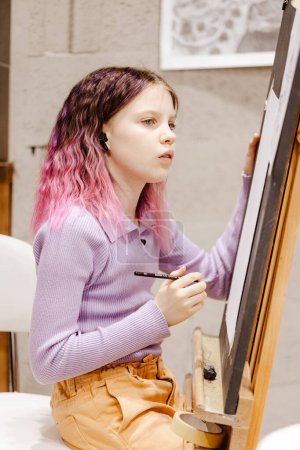 Foto de Chica 11 años de edad artesana están pintando sobre lienzo en el estudio de pie delante del caballete. Retrato de una niña pintando durante una clase de arte - Imagen libre de derechos