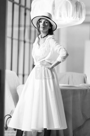 Photo for Elegant fashionable woman wearing summer white dress, straw hat, posing in stylish boho interior. Copy, empty space for text - Royalty Free Image