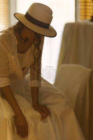 Téléchargez les photos : Élégante femme à la mode portant une robe blanche d'été, un chapeau de paille, posant dans un intérieur boho élégant. Copier, espace vide pour le texte - en image libre de droit
