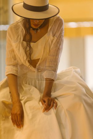Foto de Elegante mujer de moda con vestido blanco de verano, sombrero de paja, posando en un elegante interior boho. Copiar, espacio vacío para texto - Imagen libre de derechos