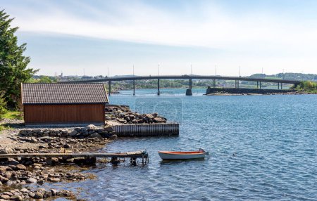 Foto de Rocky beach with a short wooden jetty and a small fishing boat near Hafrsfjord bridge, Tananger, Norway, May 2018 - Imagen libre de derechos