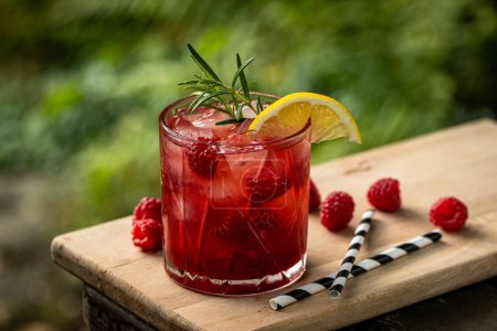 Raspberry homemade lemonade in a glass with fruit and rosemary decoration on wooden table in nature