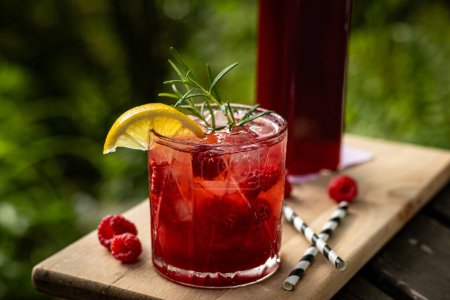 Raspberry homemade lemonade in a bottle and in a glass with fruit and rosemary decoration on wooden table in nature