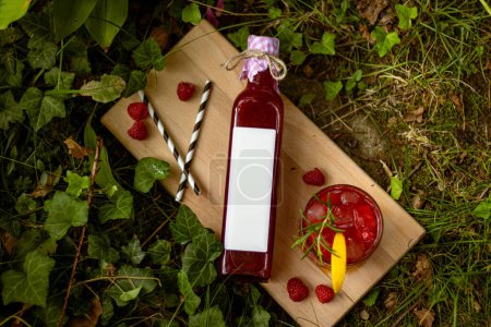 Raspberry homemade lemonade in a bottle with blank label and in a glass with fruit and rosemary decoration on wooden plater in nature