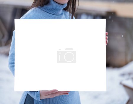 Beautiful woman holds a blank white card. Brunette girl while snow outdoors. Art mockup