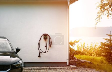 Photo for Plug holder for wall mount EV charger. Charging station with cable at wall box mounted to white house wall. Sustainable future concept. Electric cars. - Royalty Free Image