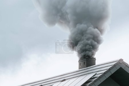 Ein modernes Haus emittiert im Winter schwarzen Rauch aus seinem Schornstein, der Umweltverschmutzung und globale Erwärmung verursacht. Schwarzer Rauch aus dem Schornstein eines Hauses auf dem Land zeigt die Auswirkungen der Heizung.