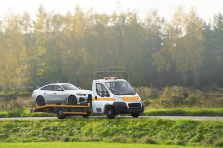 Roadside assistance transports a car with a breakdown on the road. Rollback tow truck. A tow truck delivers a failed vehicle. Roadside assistance helps a driver in trouble.