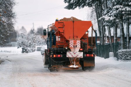 Téléchargez les photos : Un camion de chasse-neige applique du sel et du sable pendant que le camion travaille au nettoyage de la route enneigée. Comment le sel et le sable endommagent les parties métalliques d'un camion labourant la neige sur une route d'hiver - en image libre de droit