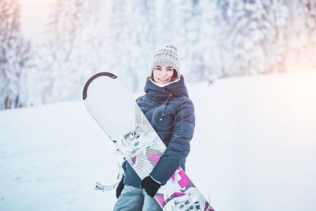 A beautiful brunette woman with a snowboard jumps on a white slope. She is enjoying the winter sport and the nature. The background is a gorgeous view of the snow-covered pines.