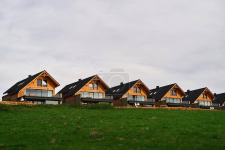 Modern wooden houses on a green hill with a stunning view of the sky and the fields. A row of modern wooden houses and green grass foreground.