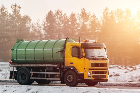 Téléchargez les photos : Un camion septique jaune avec un pétrolier vert se déplace sur une route enneigée au milieu des arbres alors que le soleil se couche et projette une lueur chaude. Camion à vide en Europe. - en image libre de droit