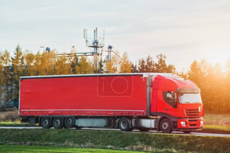 Foto de Un camión rojo transporta mercancías a través de un paisaje pintoresco iluminado por el tono dorado del sol poniente. Transporte de camiones. - Imagen libre de derechos