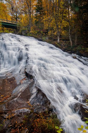 Rushing waterfall in northeast Vermont and fall foliage with highway overpass beyond.