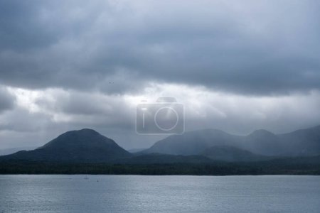 Photo for Dark scene with sunlight pushing throguh clouds with rain, hills and sea in Alaska - Royalty Free Image